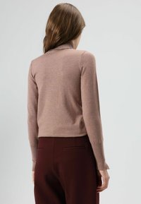 Gebreide lichtbruine trui met een hoge kraag en lange mouwen, voorzien van een zachte textuur. Draagt een strakke bordeauxrode broek.