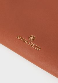 Hnědá umělá kůže s texturou, na které je vyražený text "ANNA FIELD" v zlaté barvě, s dekorativním emblémem nad písmeny.