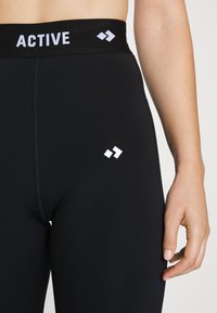 Close-up of a person wearing black leggings with "ACTIVE" text and two white diamond logos on the waistband and thigh, hand resting by side.