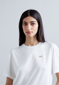 Tricou alb din bumbac cu un croi lejer, mâneci scurte și un mic logo auriu pe piept. Textură netedă și design minimalist.