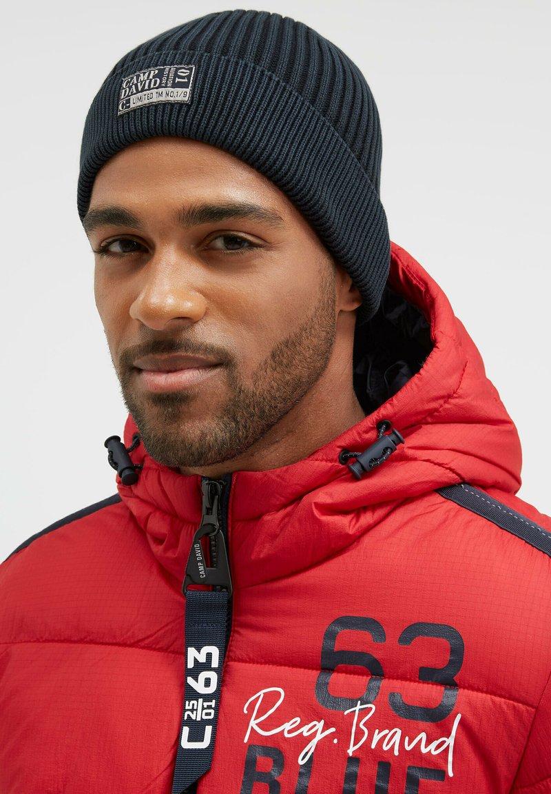 Red padded jacket with a navy blue trim and black hardware, featuring a high collar, adjustable toggles, and a logo patch on the beanie.