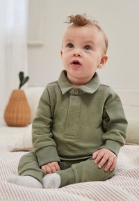 Zaļš toddler stila komplekts ar garām piedurknēm, fakturētu audumu un pogājamu apkakli, sēžot uz svītrainas virsmas, kopā ar pelēkām zeķēm.