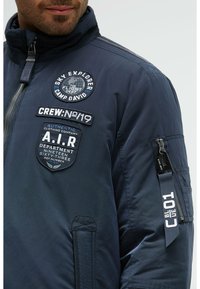 Marineblaue Jacke mit hohem Kragen, ausgestattet mit mehreren Patches, Reißverschlüssen und einem strukturierten Material. Inklusive seitlicher Taschen und einem markierten Riemen-Detail.