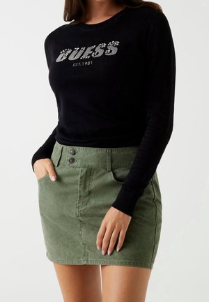 Kobieta ubrana w czarną bluzkę z długim rękawem z napisem "GUESS EST. 1981" oraz zieloną mini spódnicę na guziki, częściowo trzymająca ręce w kieszeniach.