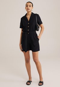 Crni kombinezon kratkih rukava s ovratnikom, zlatnim gumbima, pojasom u struku i prednjim džepovima, uparen s crnim sandalama s čvorom.
