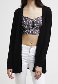 Černý pletený kardigan s otevřeným předním dílem a žebrovanými manžetami, nošený přes vzorovaný crop top, doplněný bílými těsnými džíny.