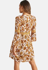 Robe florale blanche avec des motifs botaniques orange et jaune, dotée de longues manches évasées et d'un corsage ajusté qui s'évase à la taille.