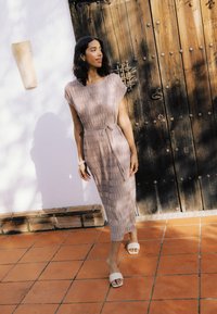 Femme en robe midi rose clair à manches courtes et sandales blanches, debout sur des carreaux en terre cuite près d'une porte en bois et d'un mur blanc sous le soleil.