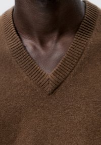 Gros plan sur le haut de la poitrine et le cou d'une personne vêtue d'un pull en maille brun à col en V.