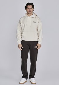 Beige hoodie met trekkoord en voorkantzak, gecombineerd met donkerbruine joggingbroek met een logo. Sneakers zijn wit met een textuur op de zool.