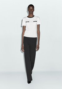 Tall woman wearing a white short-sleeve top with black trim and black wide-leg pants, walking forward against a plain light background.