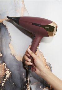 Main tenant une sèche-cheveux ghd maroon et or avec des boutons, sur un fond en marbre blanc et foncé.