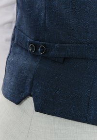 Granatowa kamizelka w wzór jodełki z dwoma czarnymi guzikami na talii, charakteryzująca się eleganckim krojem i teksturowanym materiałem. Akcenty w jaśniejszym odcieniu niebieskiego.