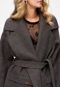 Bruine visgraatjas met een brede kraag en riem. Voorzien van grote zakken en een gestructureerde stof. Gelaagd over een zwarte sheer top.