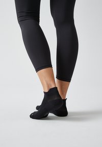 Schwarze sportliche Leggings mit einem figurbetonten Design haben eine glatte Textur und werden mit kurzen schwarzen Socken kombiniert, die am Knöchel ein Netzdetail aufweisen.