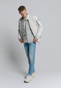 Lichtgrijze gewatteerde bomberjack, grijs gelaagd shirt, blauwe jeans en witte sneakers met beige accenten. Eenvoudig ontwerp met ritsen.