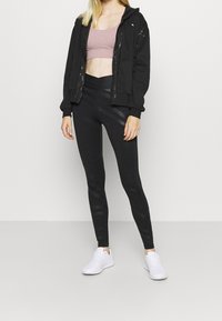 Haut à zip noir, soutien-gorge de sport rose et leggings noirs à motifs, complétés par des chaussures de sport blanches. Textures lisses et design ajusté.