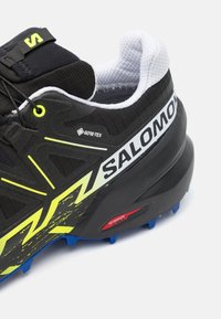 Schwarzer Laufschuh mit strukturiertem Obermaterial, neongelben Akzenten, GORE-TEX-Logo und einer blauen, genoppten Außensohle für hervorragenden Halt.