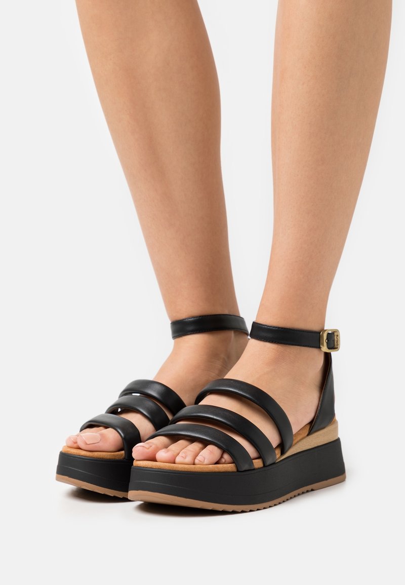 Pieds portant des sandales plateformes noires avec trois larges brides à l'avant et une bride de cheville avec une boucle dorée, sur fond blanc.