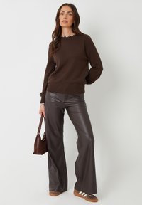 Brauner Pullover mit geripptem Saum und weißem gewelltem Halsauschnitt, kombiniert mit hochtaillierten braunen Kunstleder-Hosen mit Schlag und braunen Sneakers.