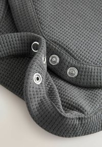 Graues Strickoberteil mit strukturiertem Muster, ausgestattet mit silbernen Druckknöpfen am Kragen und einem gerippten Bündchen für zusätzliche Details.