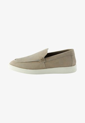 Des chaussures slip-on beige avec une tige texturée, un orteil arrondi, et une semelle en caoutchouc blanche, présentant une finition lisse et des détails de couture minimaux.