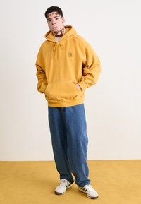 Haut en coton jaune avec une poche frontale et une capuche à cordon, associé à un jean en denim bleu et des baskets blanches. Design minimaliste.