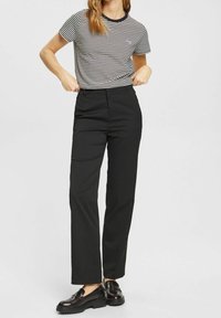 Gestreiftes Schwarz-Weiß-T-Shirt mit rundem Halsausschnitt, kombiniert mit hochgeschnittenen schwarzen Hosen und glänzenden schwarzen Loafers.