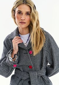 Žena s dlhými blond vlasmi nosí čierno-biely kabát so vzorom kĺboťa a červenými gombíkmi, modrý náhrdelník z korálok a bielu košeľu.