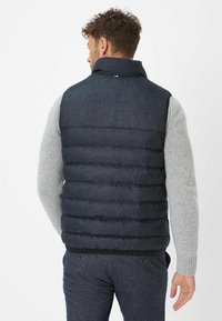 Dunkelblaues Steppweste mit hohem Kragen, ausgestattet mit gesteppter Textur, horizontalen Nähten und einem schwarzen elastischen Bund. Grauer Pullover darunter.