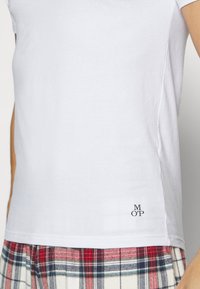 Weißes kurzärmliges T-Shirt aus Baumwolle mit einem kleinen schwarzen "MOP"-Logo am Saum, kombiniert mit karierten Hosen in Rot, Blau und Weiß.