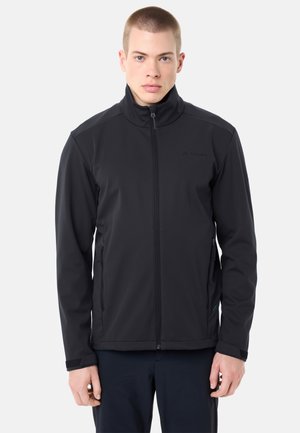 Giovane uomo con capelli corti che indossa una giacca nera con cerniera e pantaloni scuri, in piedi contro uno sfondo chiaro e uniforme.