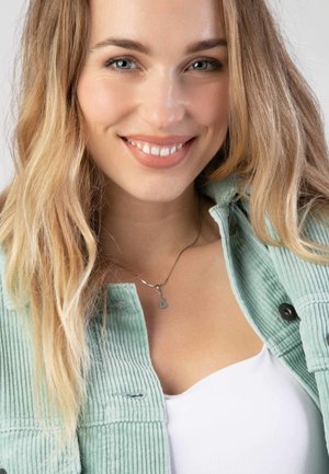 Vrouw die glimlacht, gekleed in een lichtgroene corduroyjas en witte top, met een zilveren gitaarhanger als ketting. Haar in losse golven.