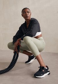 Felpa nera corta con una vestibilità ampia, leggings verde chiaro e scarpe da ginnastica nere con accenti arancioni, mentre afferra una corda nera.