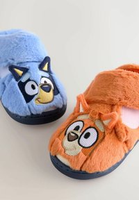 Chaussons bleu et orange duveteux, ornés de visages de personnages de dessins animés. Matériau textil doux, semelles noires et détails brodés sur chaque chausson.