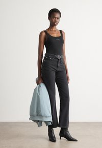 Femme portant un débardeur noir, un jean noir taille haute et des bottines noires, tenant une veste bleu clair pliée dans sa main gauche.
