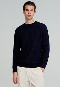 Granatowy sweter w prążki wykonany z miękkiego materiału, z okrągłym dekoltem i długimi rękawami, w połączeniu z jasnobeżowymi spodniami.