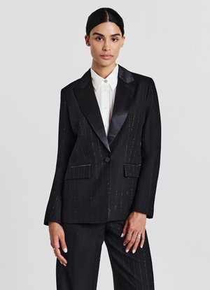 Schwarzer Blazer mit Nadelstreifen und Satinrevers, zwei Außentaschen und einem einzelnen Knopfverschluss. Kombiniert mit einem weißen Hemd mit Kragen darunter.