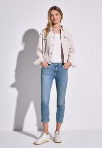 Chaqueta corta de color beige claro sobre una camiseta blanca, combinada con unos vaqueros ajustados azules claros y zapatillas blancas. Diseño sencillo con acentos sutiles.