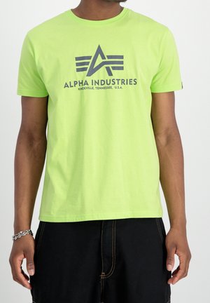 Mann trägt limonengrünes Alpha Industries T-Shirt mit Logo und Schriftzug, kombiniert mit schwarzen Hosen vor schlichtem weißen Hintergrund.