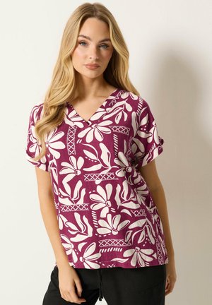 Blondynka w krótkim, fioletowym bluzce w białe kwiatowe wzory i czarnych spodniach, stojąca na jednolitym tle.