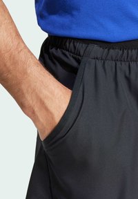 Black athletic shorts featuring a textured fabric, elastic waistband, and side pockets. A hand rests in the left pocket.