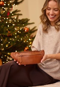 Usměvavá žena v béžovém svetru drží hnědou koženou zipovací tašku, sedí vedle ozdobeného vánočního stromečku s světýlky a oranžovými ozdobami.