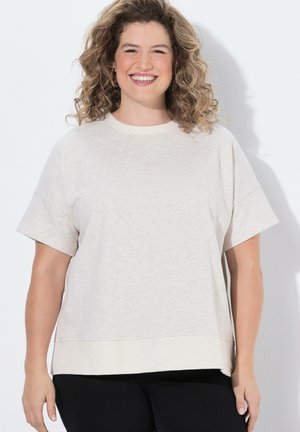 Smilende kvinde med krøllet hår iført en beige kortærmet top og sorte bukser, stående mod en hvid baggrund.