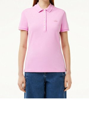 Femme portant un polo rose clair à manches courtes avec des boutons et un jean bleu, debout, les bras détendus le long du corps.
