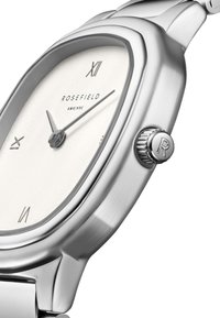 Reloj de acero inoxidable plateado con una caja rectangular, esfera blanca y marcadores de hora minimalistas en negro. Presenta una corona texturizada con un detalle de "R".