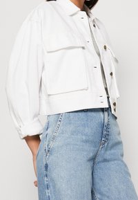 Giacca di jeans bianca corta con chiusura a bottoni, due tasche frontali e una vestibilità comoda. Abbinata a jeans a vita alta di colore azzurro chiaro.