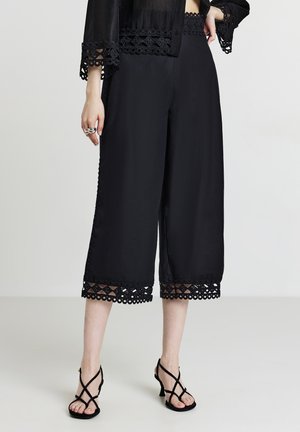 Schwarze, hochgeschnittene Hosen mit Spitzenbesatz am Saum, die eine glatte Textur und einen lockeren Schnitt aufweisen. Kombiniert mit schwarzen Riemchen-High-Heels.