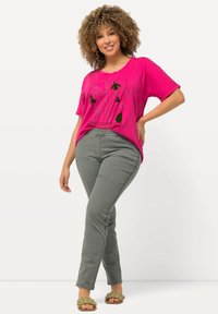 Růžové grafické tričko s černým květinovým vzorem, spárované se šedými slim-fit kalhotami. Modelka nosí zelené pletené sandály. Jednoduché pozadí.