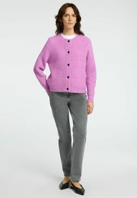 Rosa Wollcardigan mit rundem Ausschnitt, Knopfverschluss und langen Ärmeln. Kombiniert mit grauen geraden Hosen und schwarzen Ballerinas.